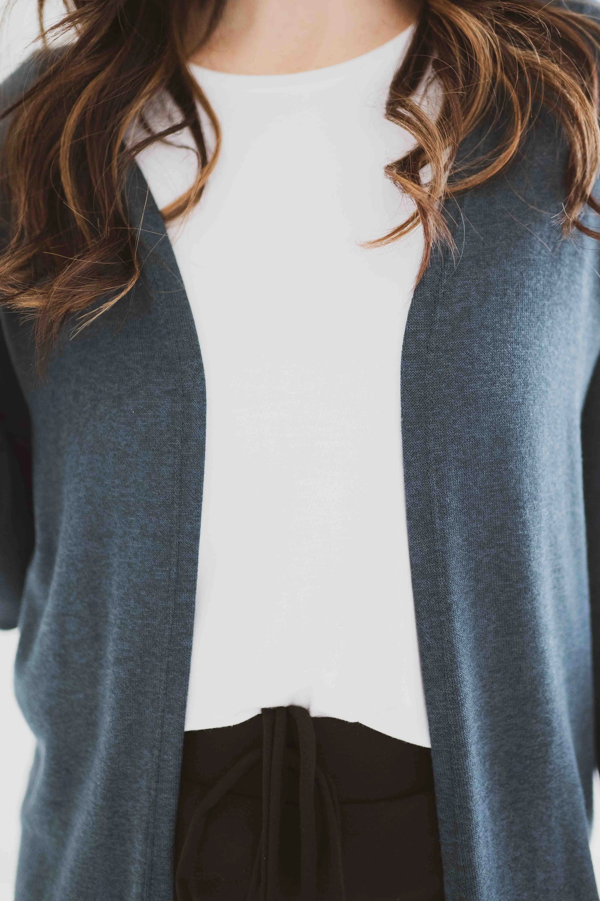 Close up of a woman wearing the Carolina Cardigan by Ang Hill in Juniper, an open cardigan with long sleeves. She is wearing it with black pants and a white top and is standing in front of a white background. 