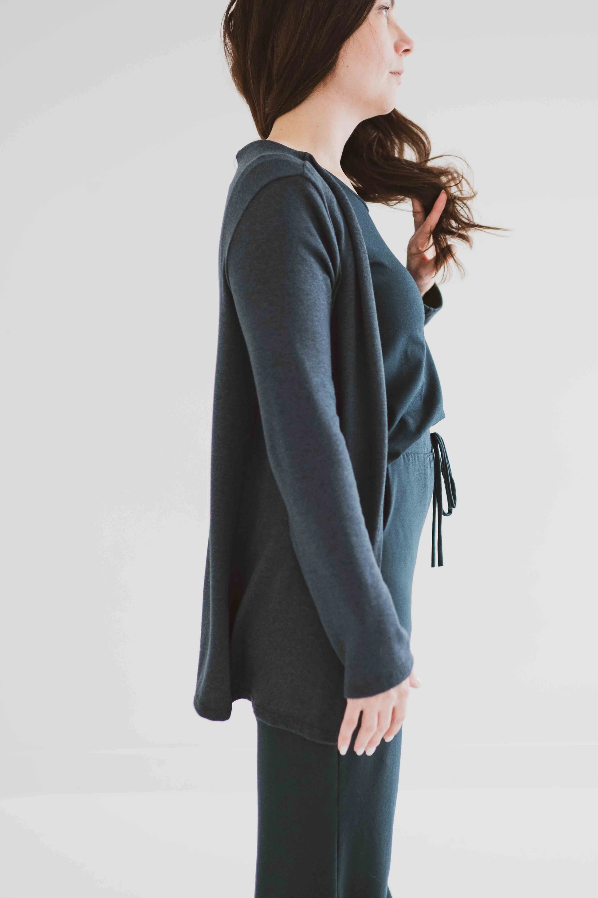 Side view of a woman wearing the Carolina Cardigan by Ang Hill in Juniper, an open cardigan with long sleeves. She is wearing it with black pants and a white top and is standing in front of a white background. 