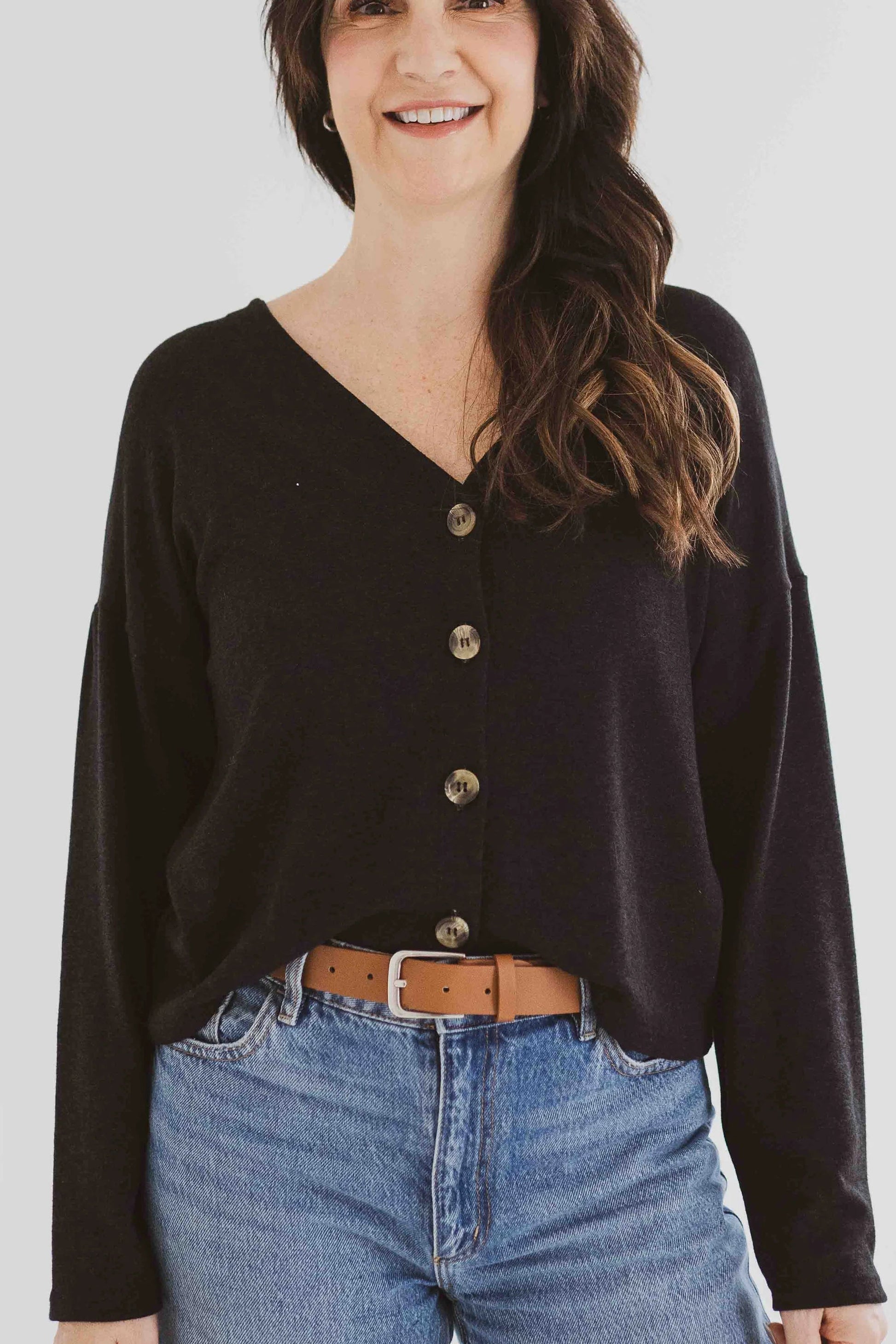 A woman wearing the Holly Cropped Cardigan by Ang Hill in Black, a v-neck cardigan with drop sleeves and tortoiseshell buttons. She is wearing it with jeans and is standing in front of a white background. 