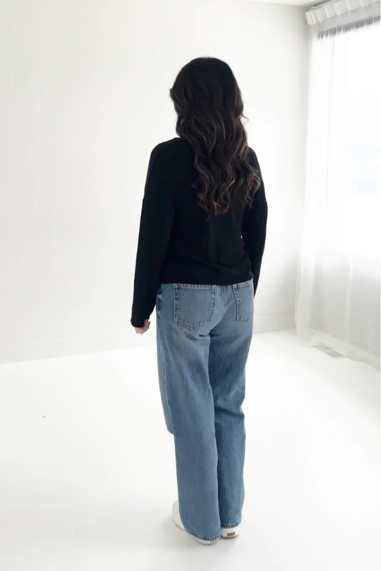 Back view of a woman wearing the Holly Cropped Cardigan by Ang Hill in Black, a v-neck cardigan with drop sleeves and tortoiseshell buttons. She is wearing it with jeans and is standing in front of a white background. 