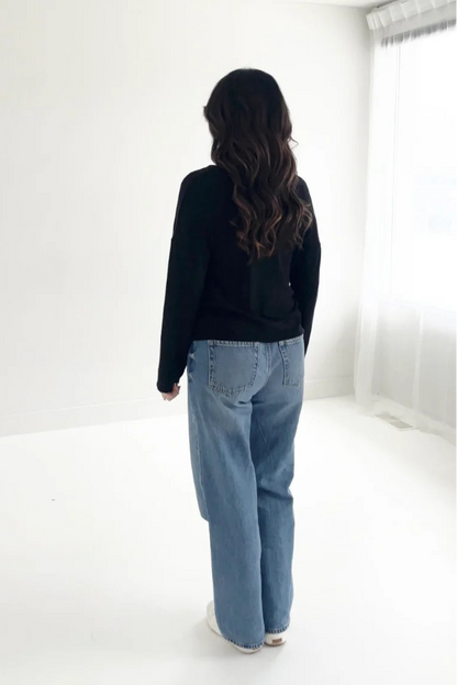 Back view of a woman wearing the Holly Cropped Cardigan by Ang Hill in Black, a v-neck cardigan with drop sleeves and tortoiseshell buttons. She is wearing it with jeans and is standing in front of a white background. 