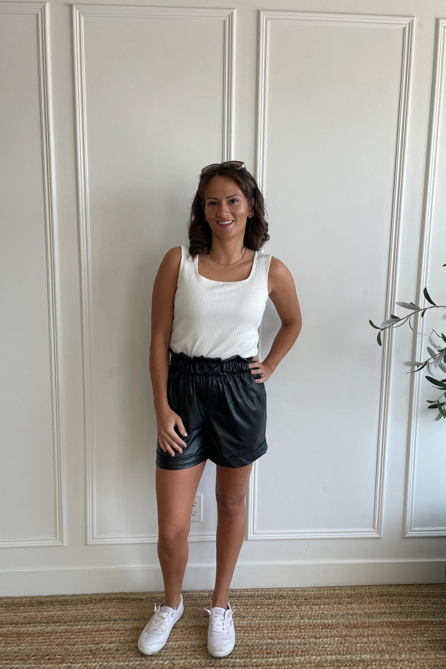 A woman wearing the Ang Hill Tracey Tank in Vanilla, with wide straps, ribbed fabric, and a square neckline. She is wearing it with black shorts and standing against a white paneled wall.