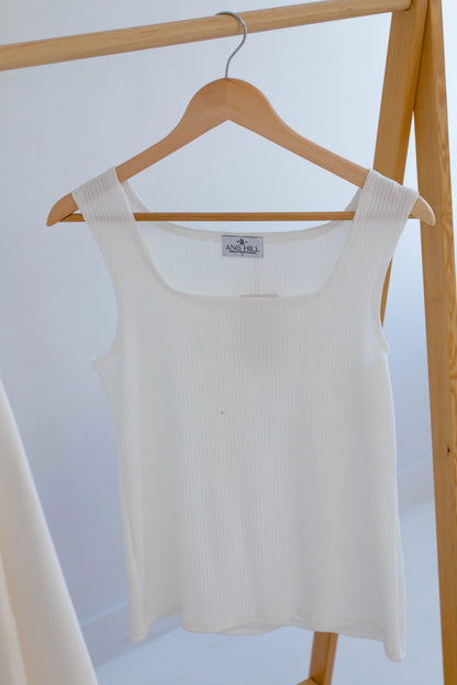 The Ang Hill Tracey Tank in Vanilla, with wide straps, ribbed fabric, and a square neckline, is shown hanging on a wooden rack. 