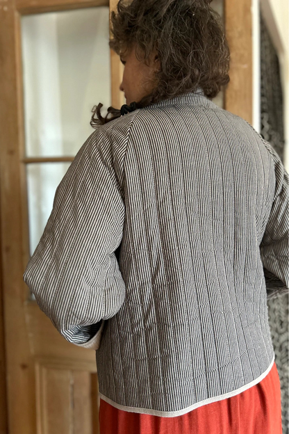 Back view of a woman wearing the Artemesia Jacket by Kazak in Taupe Stripe, a quilted jacket with snaps up the front and large patch pockets. She is wearing it over  the Lavendula Dress Paprika and standing in front of a wood and glass door. 