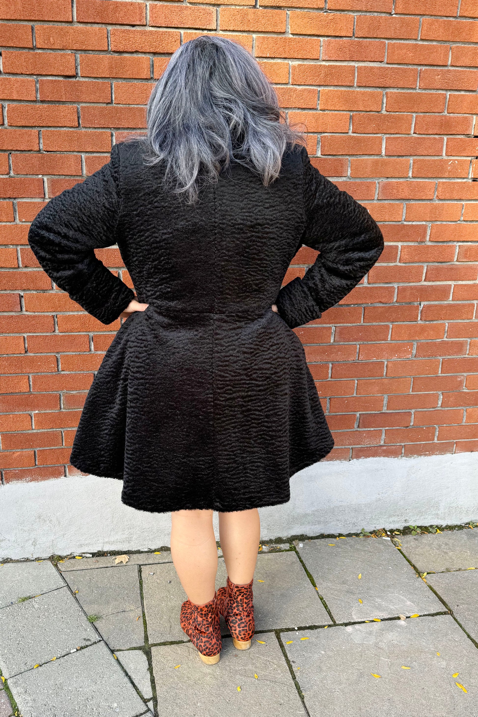 Back view of a woman wearing the Spies Jacket by Luc Fontaine,  
a fit and flare above the knee coat in boucle fabric, standing in front of a brick wall 