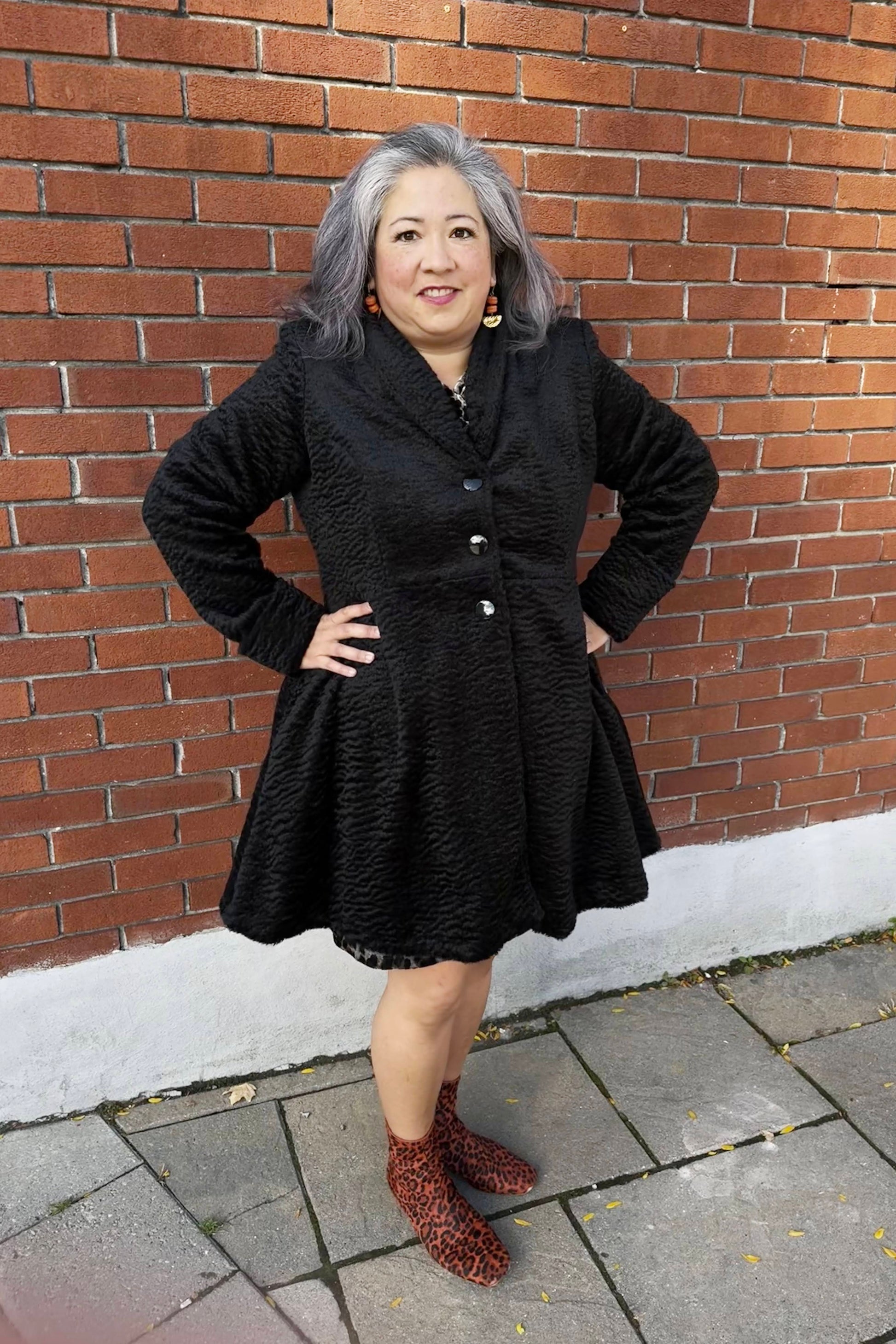 A woman wearing the Spies Jacket by Luc Fontaine,  
a fit and flare above the knee coat in boucle fabric, standing in front of a brick wall 