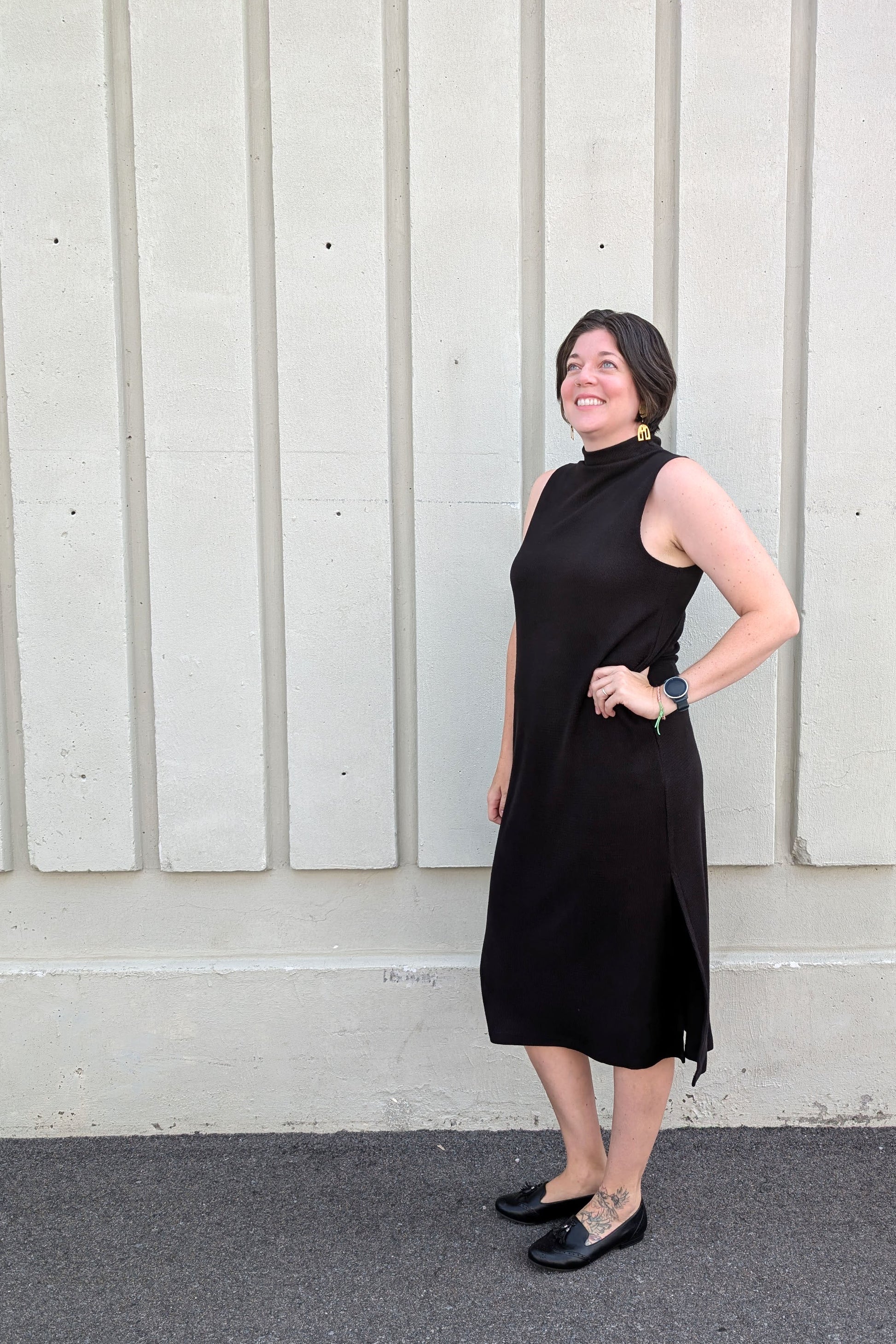 Side view of a woman wearing the Natasha Dress by Compli K in Black, a sleeveless midi-length dress with a mock turtleneck and side slits, standing outside in front of a white wall 