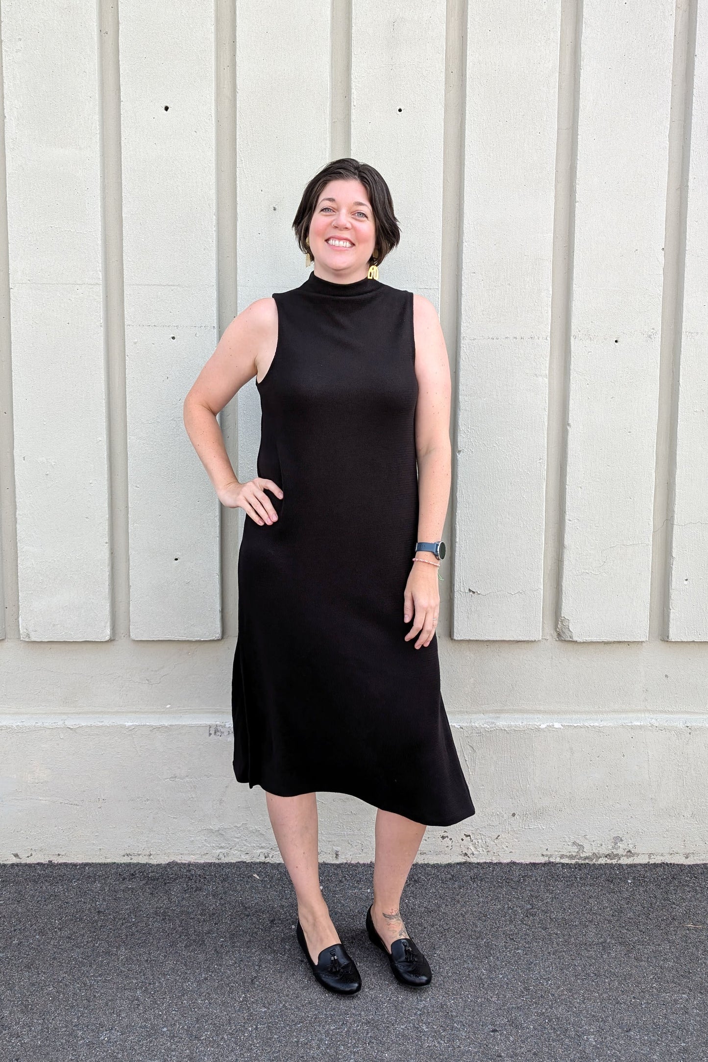 Side view of a woman wearing the Natasha Dress by Compli K in Black, a sleeveless midi-length dress with a mock turtleneck and side slits, standing outside in front of a white wall 