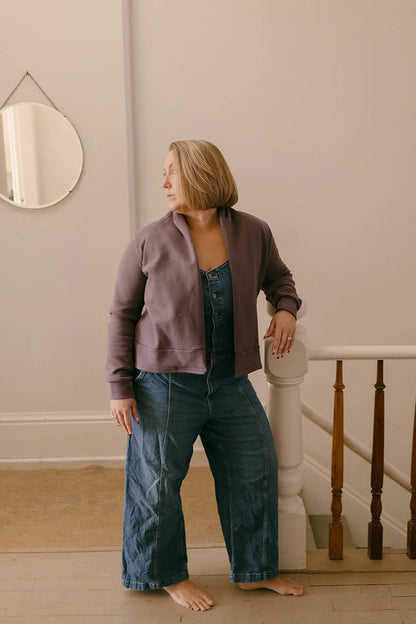 A woman wearing the Classic Spruce Cardigan by Blondie in Amethyst, a short cardigan with a subtle lapel collar, long sleeves, a wide ribbed hem. She is wearing it over denim overalls and is standing in front of a banister.