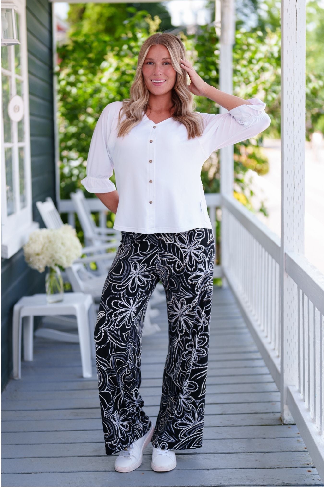 A woman wearing the Coco Palazzo Pants by Rien ne se Perd in Black and White floral, with a wide elastic waistband and wide legs. She is wearing them with a white top and standing on a wooden porch.