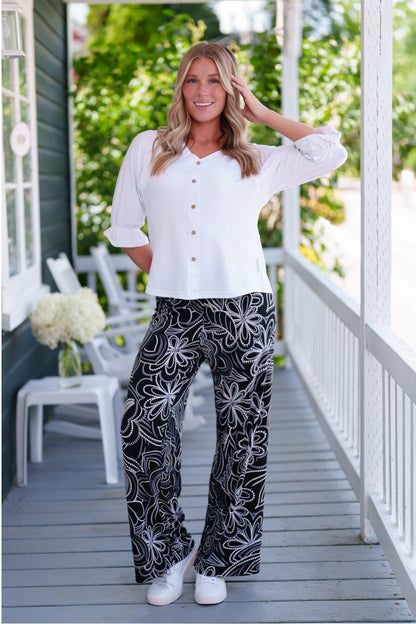 A woman wearing the Coco Palazzo Pants by Rien ne se Perd in Black and White floral, with a wide elastic waistband and wide legs. She is wearing them with a white top and standing on a wooden porch.