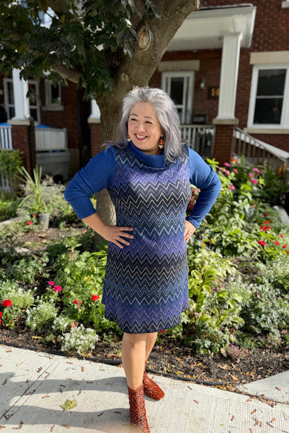 A woman wearing the Haizel Dress by Luc Fontaine in Blue, featuring a cowl neck, long sleeves, and a chevron pattern, standing in front of a garden 

