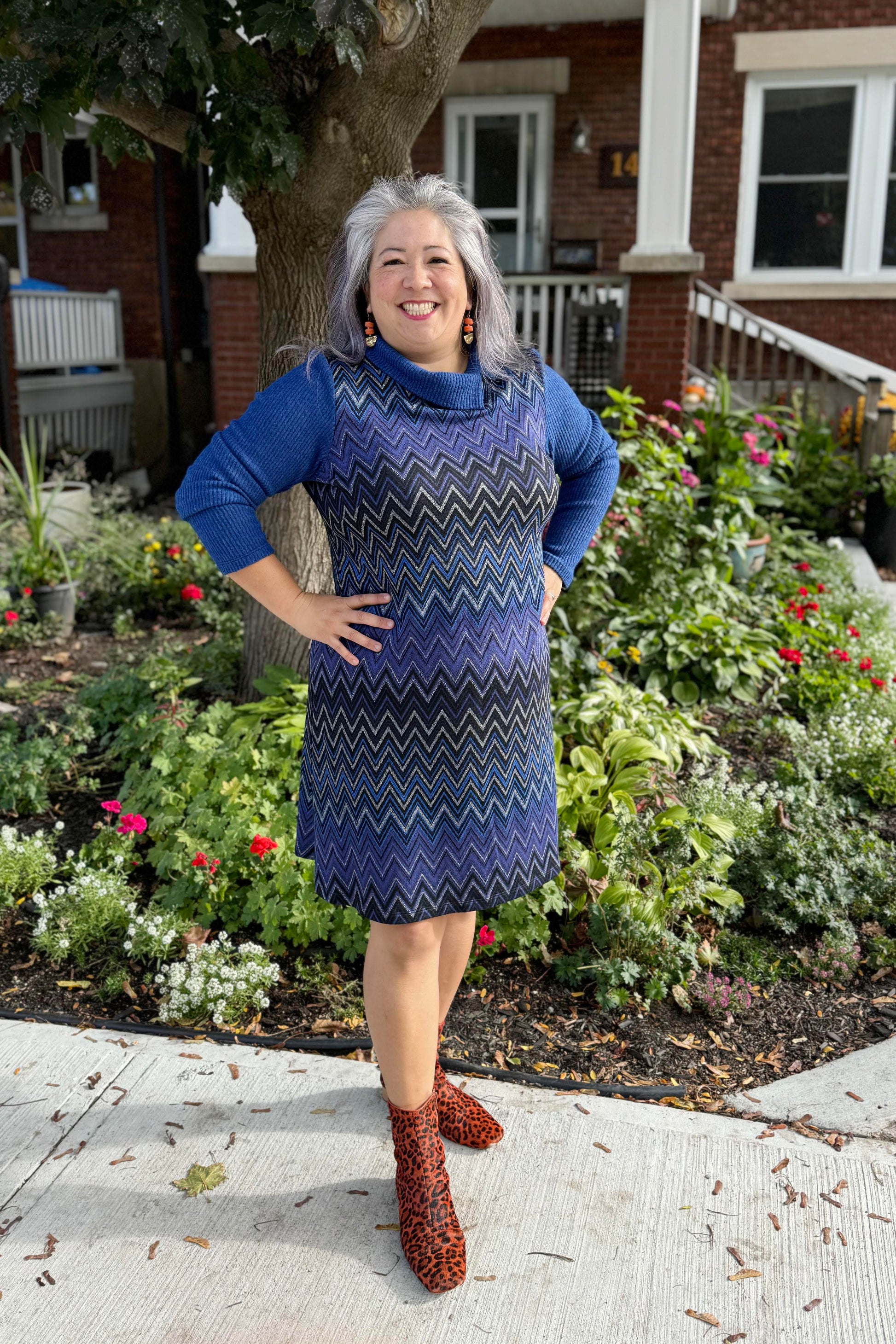 A woman wearing the Haizel Dress by Luc Fontaine in Blue, featuring a cowl neck, long sleeves, and a chevron pattern, standing in front of a garden 