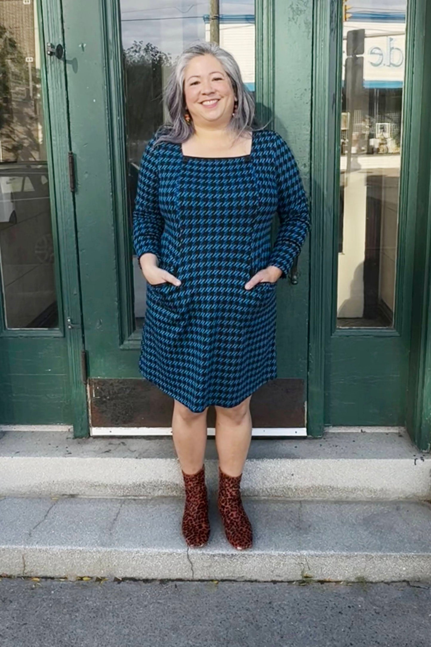 A woman wearing the Charing Dress by Luc Fontaine in Blue houndstooth, featuring a square neckline with button details, patch pockets, and a A-line shape. She is standing in front of a green doorway. 