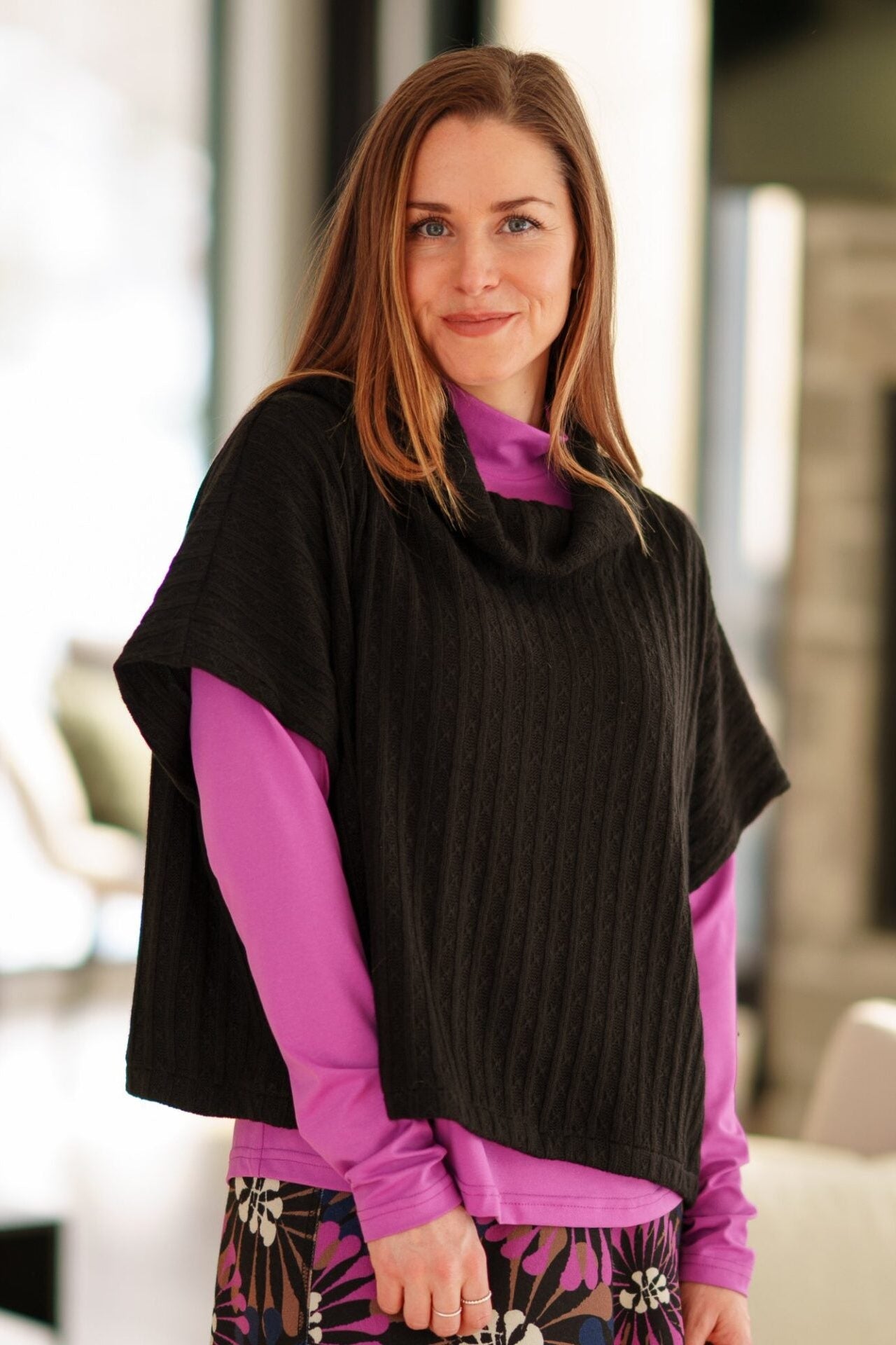 A woman wearing the Grandmont Bolero by Rien Ne Se Perd in Black,  a ribbed cowl-neck poncho. She is wearing it  over the Ste Croix Sweater in Magenta and the floral Hudson Skirt and standing in a softly lit living room with a fireplace in the background.