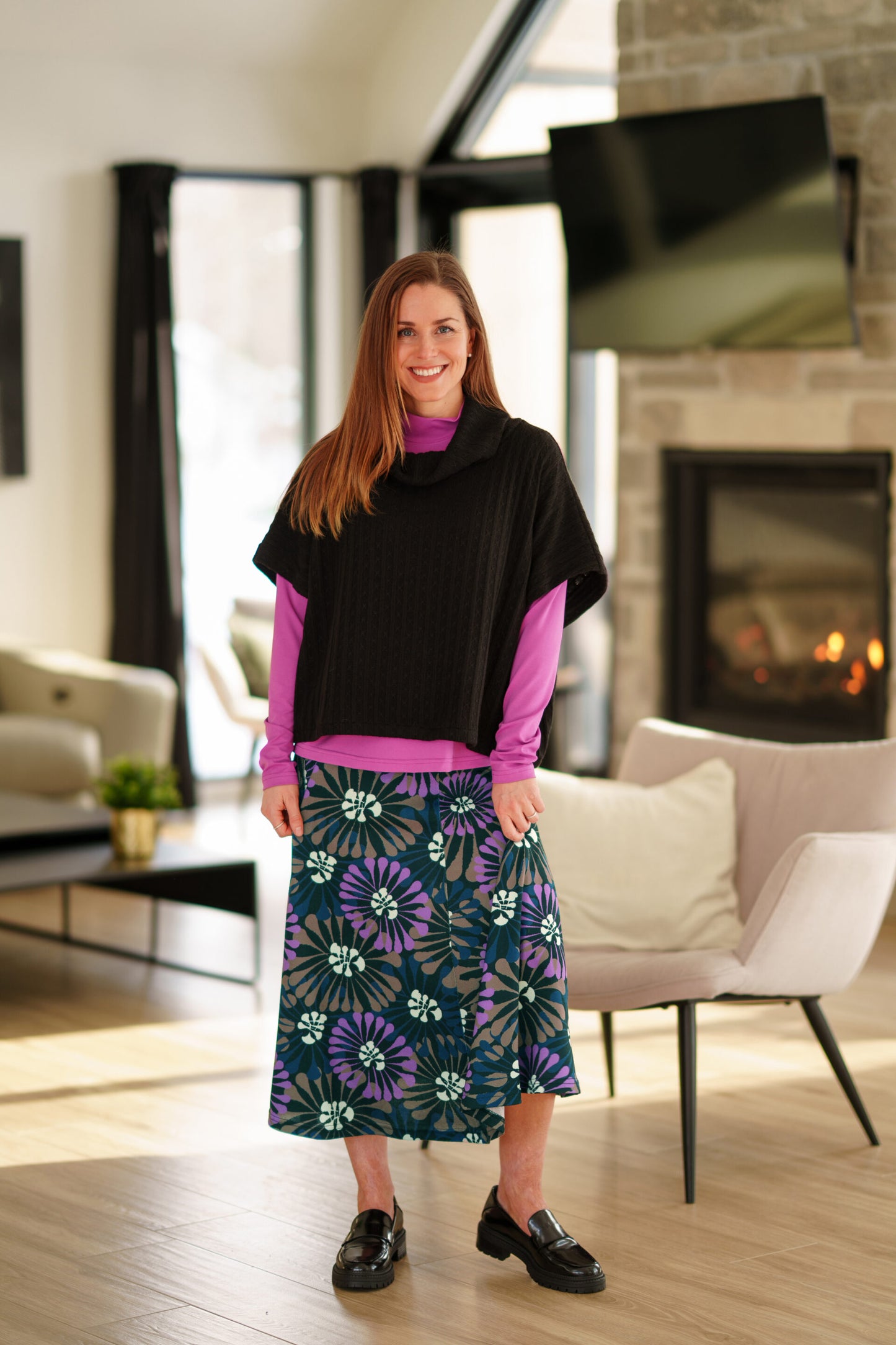 A woman wearing the Grandmont Bolero by Rien Ne Se Perd in Black,  a ribbed cowl-neck poncho. She is wearing it  over the Ste Croix Sweater in Magenta and the floral Hudson Skirt and standing in a softly lit living room with a fireplace in the background.