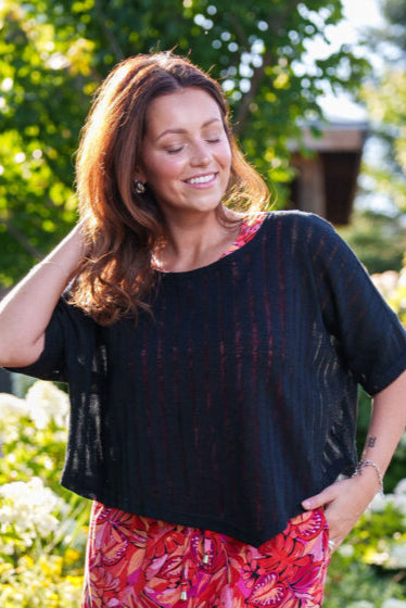 A woman wearing the Menuet Bolero- Poncho by Rien ne se Perd, a Black woven lightweight poncho. She's wearing it over a floral dress and standing outside. 