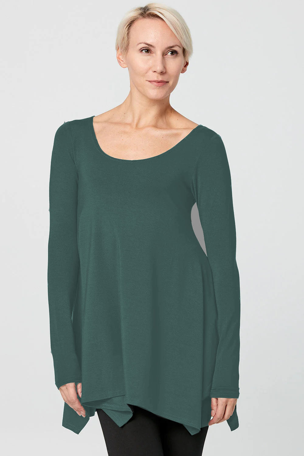 A woman wearing the Breath Top by Advika in Pine, featuring a scooped neck, long sleeves, and a swing shape with an asymmetrical hem, all in organic cotton/tencel. She is wearing it with leggings and standing in front of a white background. 