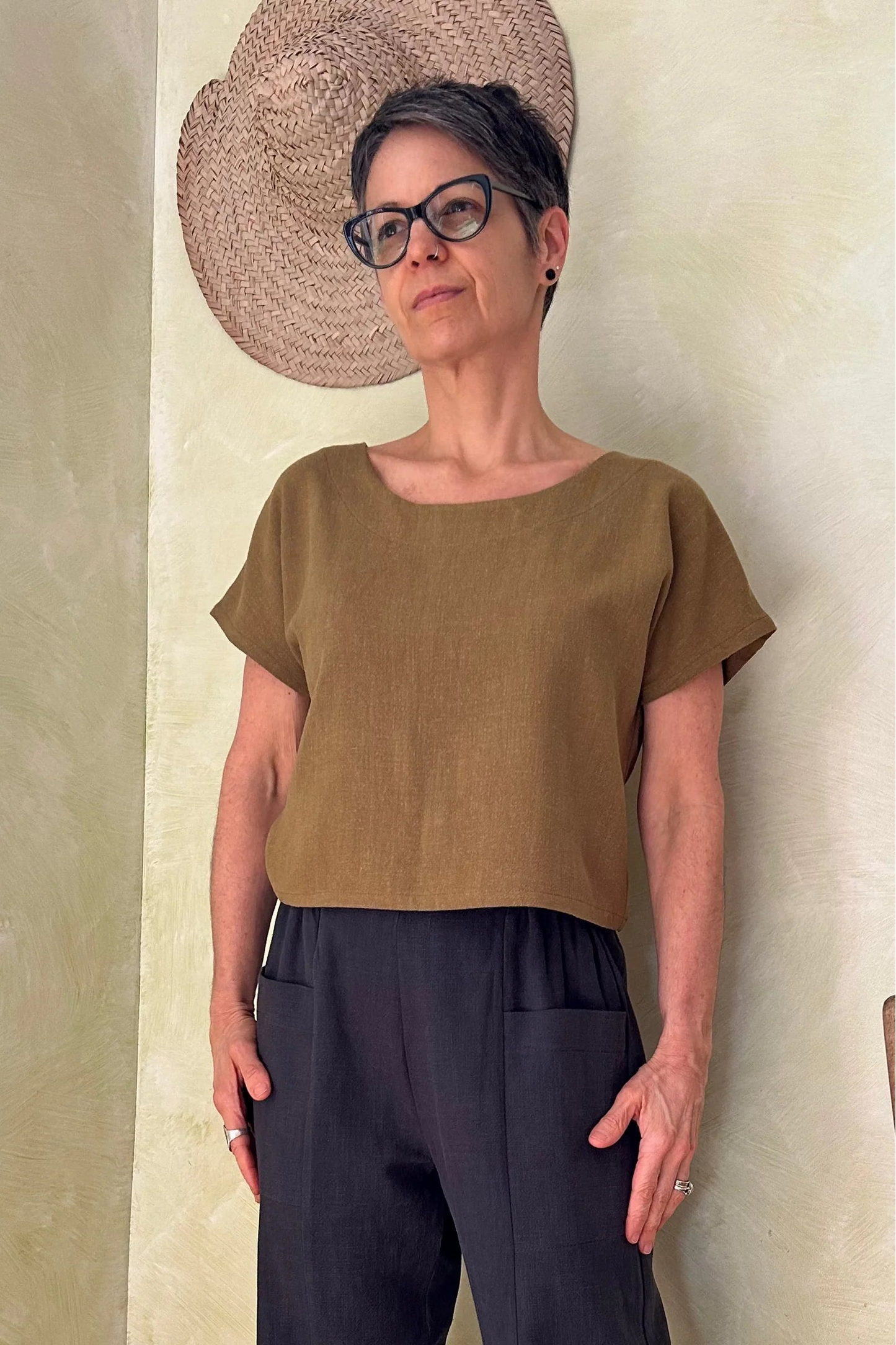 A woman wearing the Brendola Top from Kazak in Mousse, a slightly cropped short sleeved top with three brass snaps that can be worn at the back or the front. She is wearing it with the navy pants and standing in a room with hats on the wall. 
