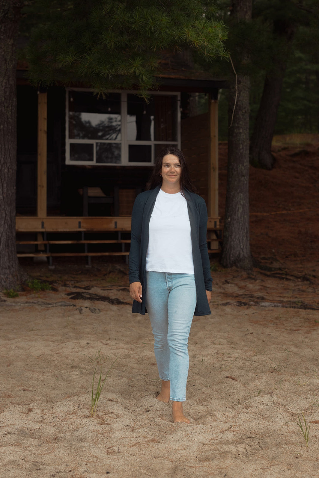 A woman wearing the Briton Cardigan from Blondie in Poppy, a mid-thigh length open cardigan with a small collar and in-seam pockets. She is wearing it with a white tee shirt and jeans and outside a cabin. 