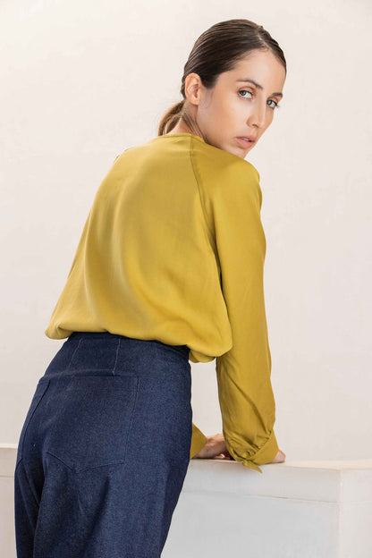 Back view of a woman wearing the Cairo Blouse by MAS in Lime, made from lyocell with raglan sleeves with gathered cuffs, and a keyhole and cord neckline. She is wearing it with the Vienna Trousers in Jeans and standing in front of a white background. 