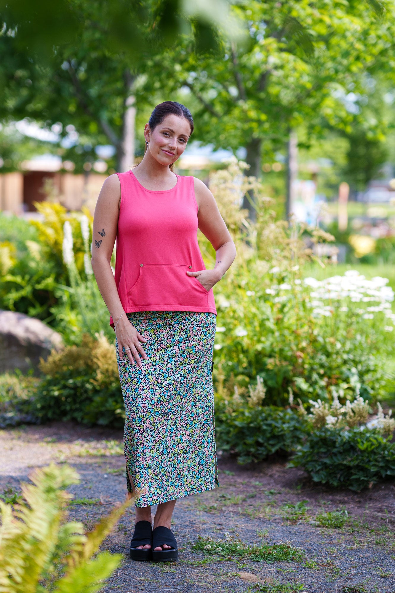 A woman wearing the Madison 7/8 Skirt by Rien ne se Perd in a Tiny Flowers pattern, featuring a straight cut with side slits and an elastic waist. She's wearing it with a coral tank top and is standing in a garden.