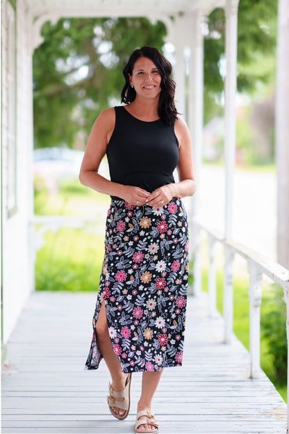 A woman wearing the Fiesta 7/8 Skirt by Rien ne se Perd in Colourful Daisies, a slightly A-line skirt with an elastic waist and side slits. She is wearing it with a black tank and standing on a wooden porch.
