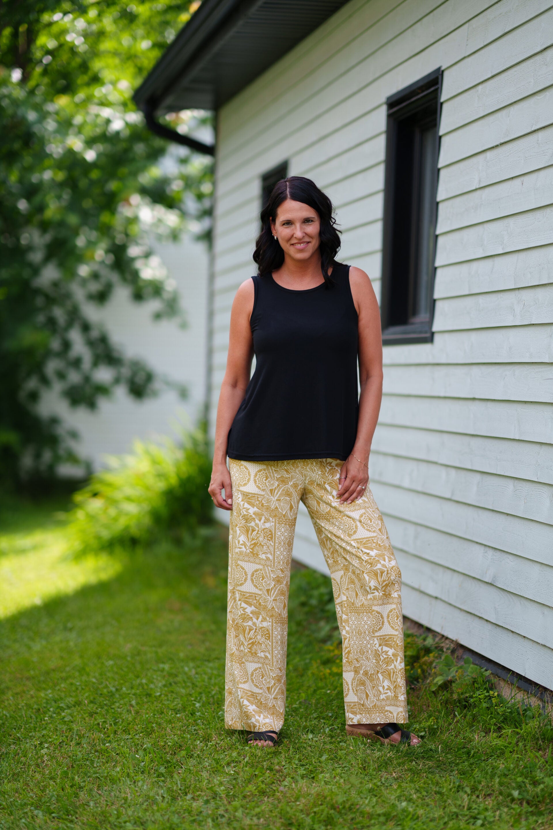 A woman wearing the Spritz Tank Top by Rien ne se Perd in Black, with wide straps and side slits. She is wearing it with  a black tank top and patterned pants and standing in front of a house.