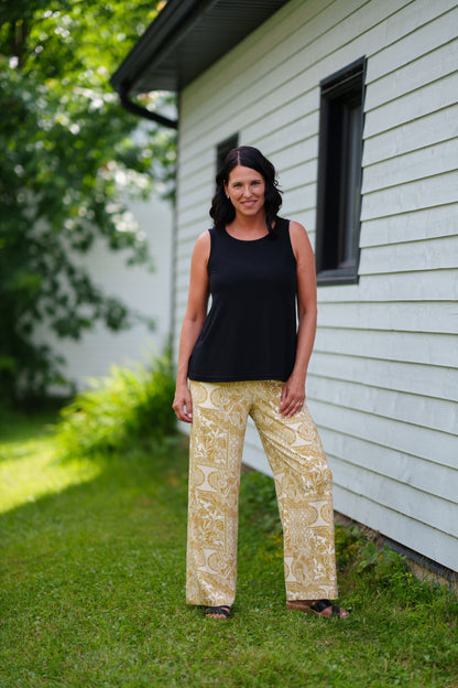 A woman wearing the Spritz Tank Top by Rien ne se Perd in Black, with wide straps and side slits. She is wearing it with  a black tank top and patterned pants and standing in front of a house.