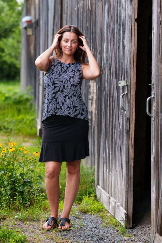 A woman wearing the Swing Cami by Rien ne se Perd in Black and White Foliage, a trapeze cut tank with a hi-low rounded hemline. She is wearing it with a black skirt and standing outside in front of a wooden building. 