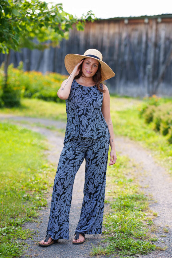 A woman wearing the Rumba Palazzo Pants in Black and White Foliage and the Swing Cami in Black and White Foliage by Rien ne se Perd, she is standing outside in front of a wooden building. 