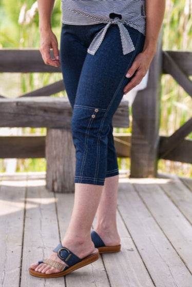 A waist-down view of a woman wearing the LIndy Capri pants from Rien ne se Perd, faux denim stretch capris with a wide pull-on waist, contrast seams at the bottom, and decorative coconut buttons. She is wearing them with a striped t-shirt and standing in front of a wooden fence. 