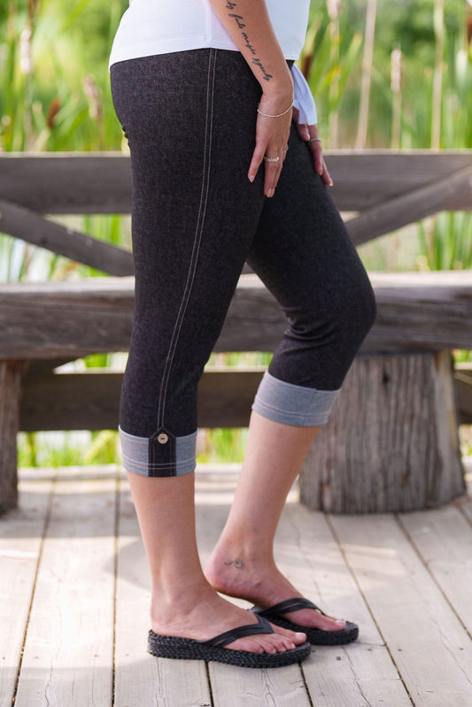 Waist-down view of a woman wearing the Miley Capri by Rien ne se Perd in Grey Denim, a pull-on capri in  stretchy fabric with a wide waistband and contrast cuffs with buttoned tab. She is wearing them with a white top and standing outside. 