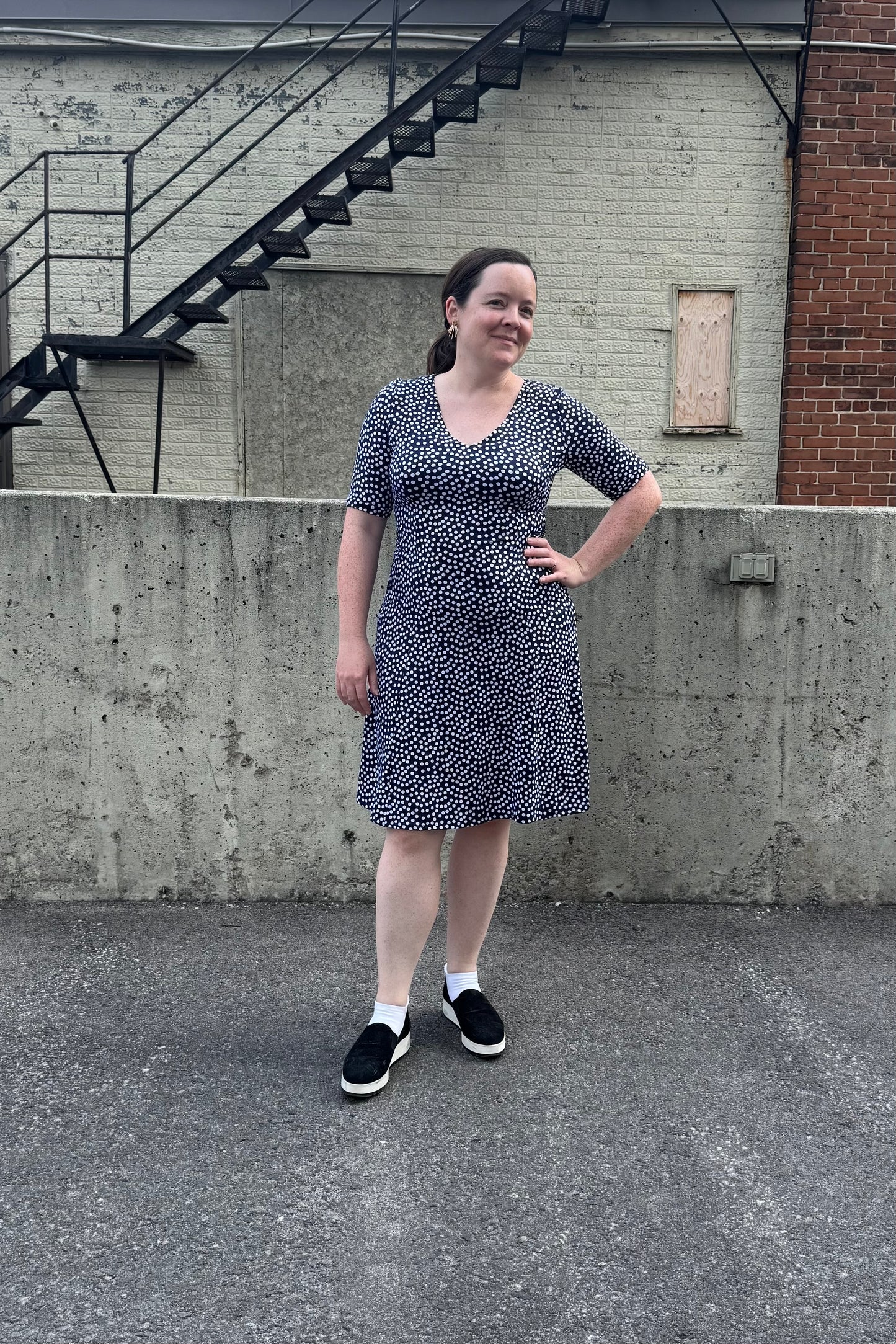 A woman wearing the Navy Polka Dot Dresss by Karkass. It has a v-neck, short sleeves, princess seams, and an above the knee hem. She is standing in front of a brick wall. 