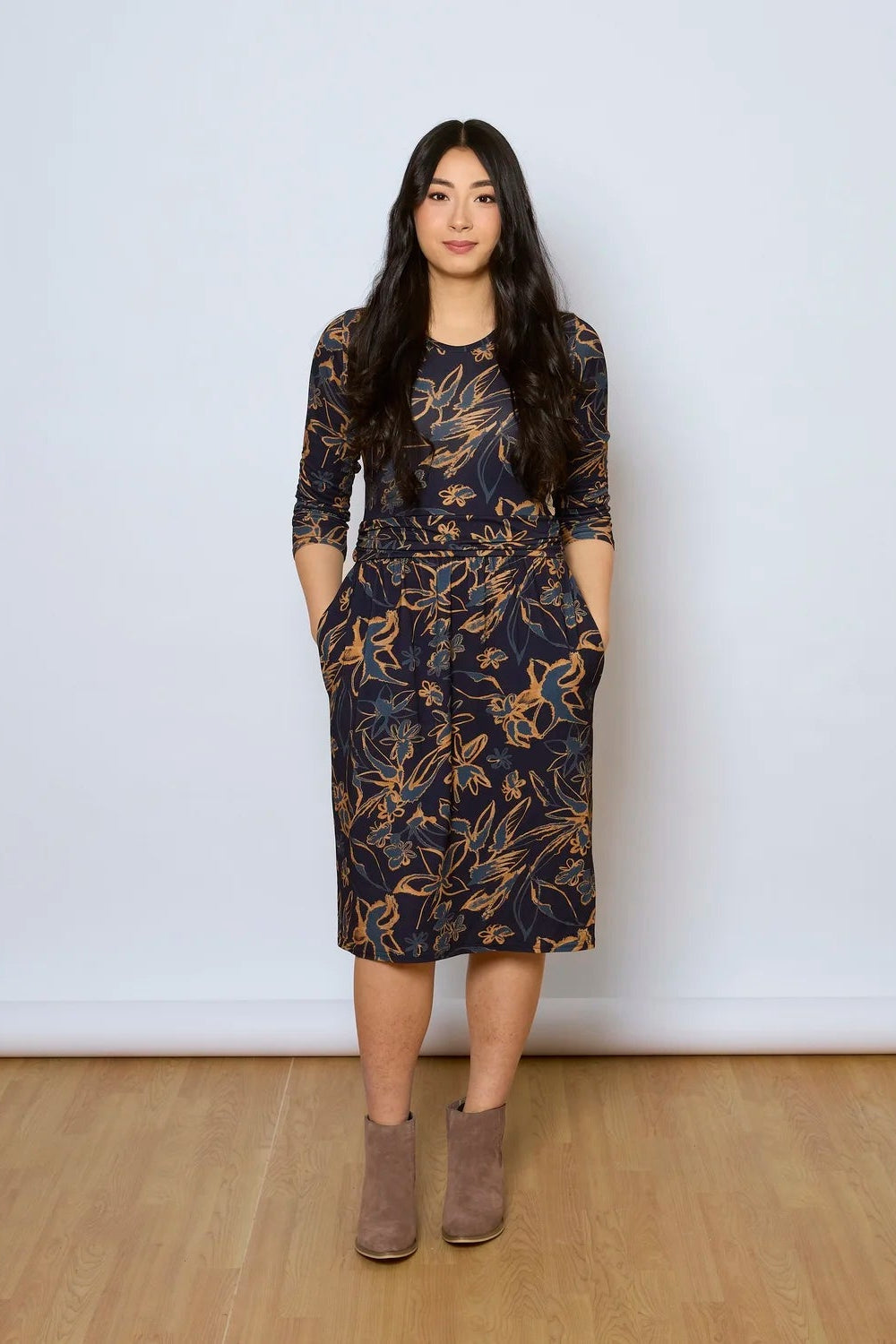 A woman wearing the Cascade Dress by Tangente in Navy Print, with a round neck, 3/4 sleeves, gathered defining waistband, and a gathered knee-length skirt. She is standing against a white wall.