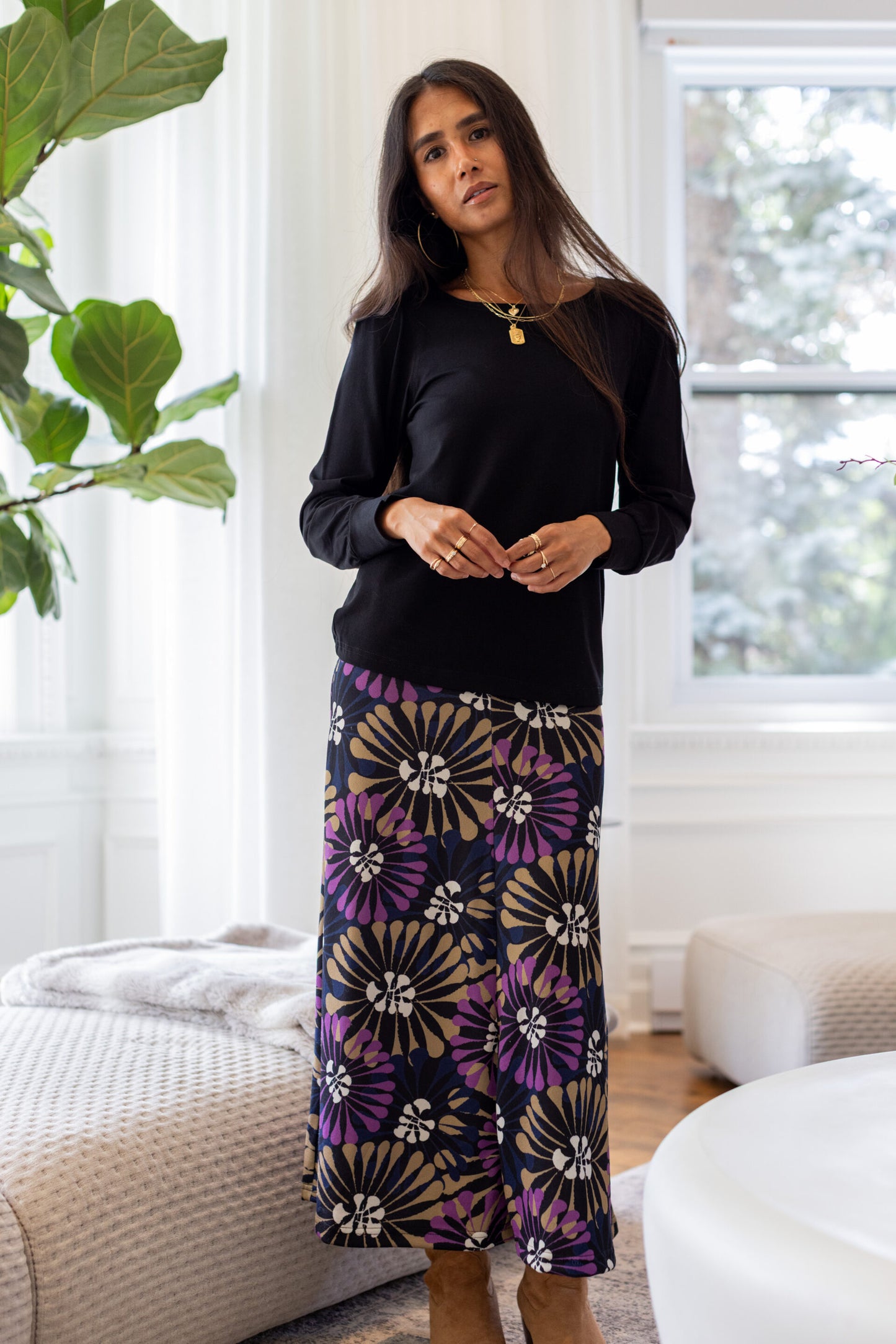 A woman standing in a white living room wearing the Becancour Top by Rien ne se Perd in Black, with a round neck and long slightly puffed sleeves with gathered cuffs. She is wearing it with the Hudson Skirt in Floral.
