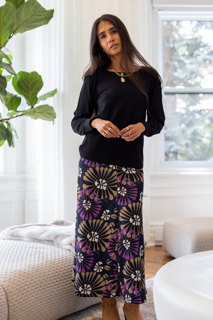 A woman standing in a white living room wearing the Becancour Top by Rien ne se Perd in Black, with a round neck and long slightly puffed sleeves with gathered cuffs. She is wearing it with the Hudson Skirt in Floral.
