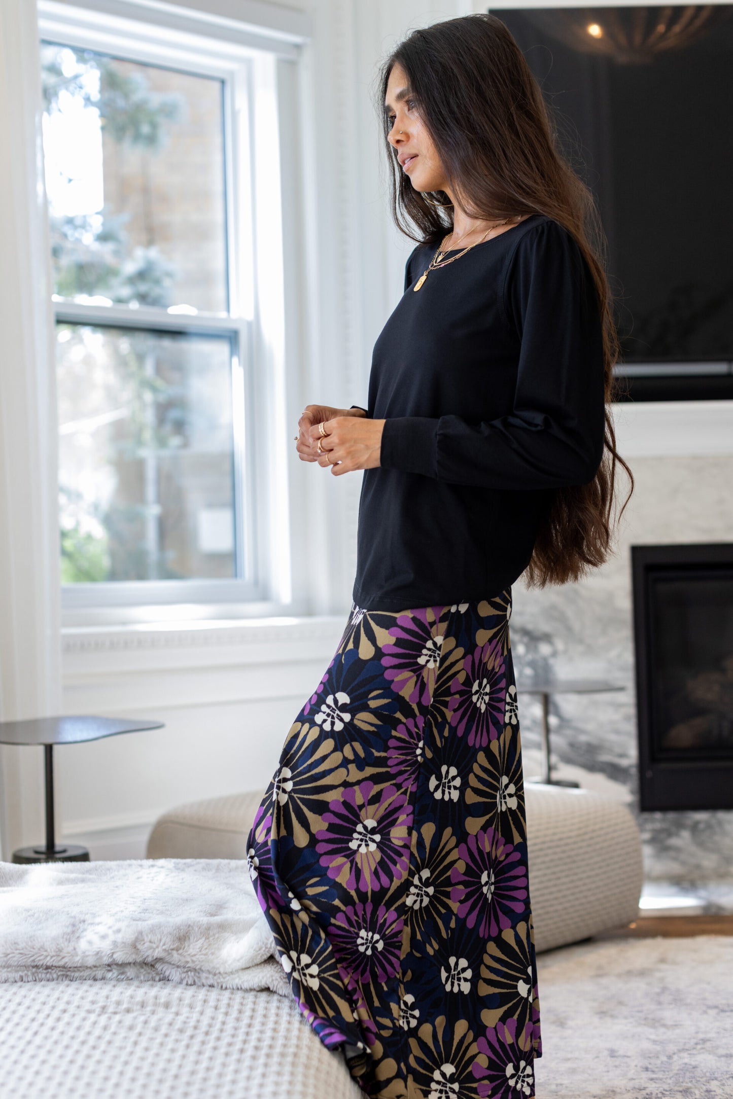 Side view of a woman standing in a white living room wearing the Hudson Skirt by Rien ne se Perd in Floral, a midi-length skirt with a bold purple, gold, and white floral burst pattern. She is wearing it with the Becancour Top in Black. 