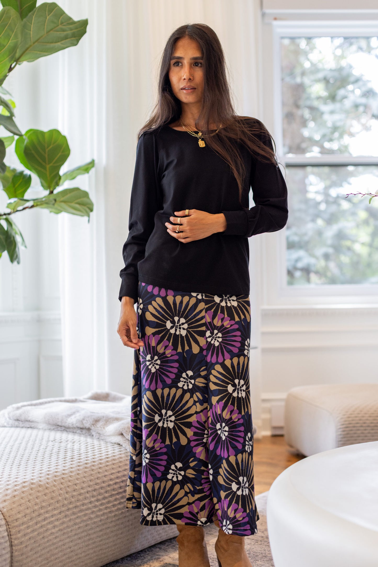 A woman stands in a white living room wearing the Hudson Skirt by Rien ne se Perd in Floral, a midi-length skirt with a bold purple, gold, and white floral burst pattern. She is wearing it with the Becancour Top in Black. 