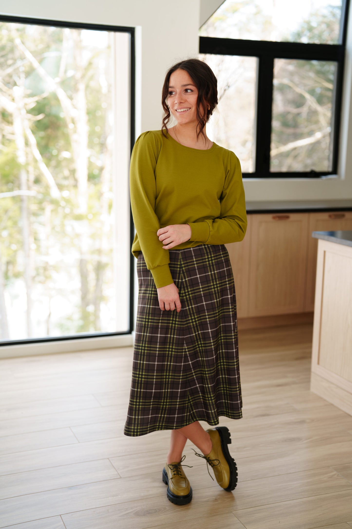 A woman wearing the Becancour Top by Rien ne se Perd in Pistachio, with a round neck and long slightly puffed sleeves with gathered cuffs. She is wearing with a plaid skirt and standing indoors in front of large windows. 
