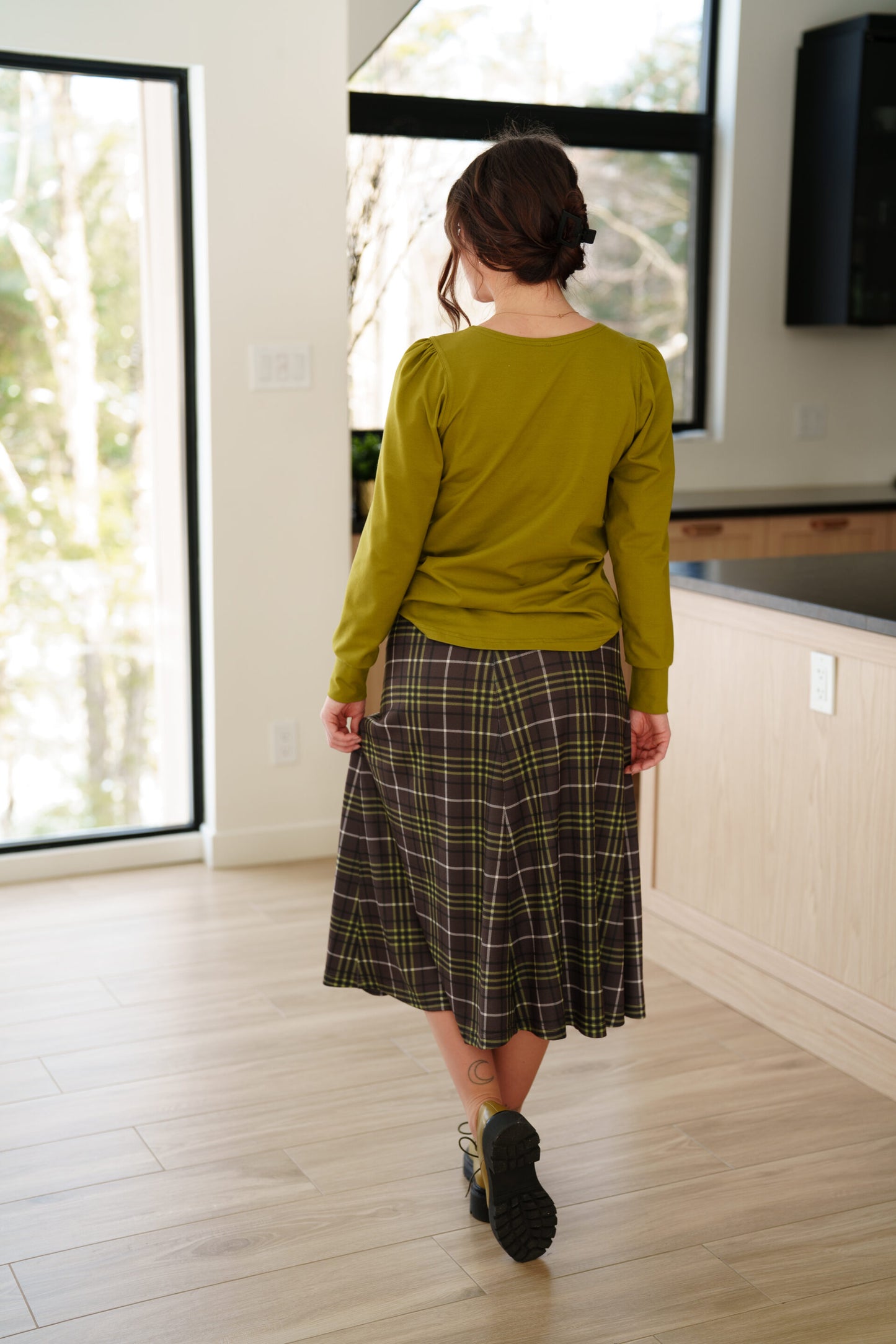 Back view of a woman wearing the Becancour Top by Rien ne se Perd in Pistachio, with a round neck and long slightly puffed sleeves with gathered cuffs. She is wearing with a plaid skirt and standing indoors in front of large windows. 
