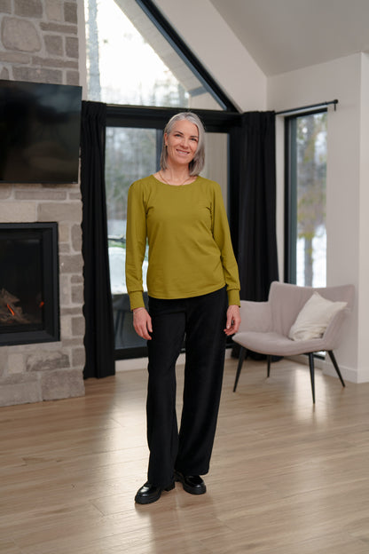 A woman wearing the Becancour Top by Rien ne se Perd in Pistachio, with a round neck and long slightly puffed sleeves with gathered cuffs. She is wearing it with black pants and standing in a living room with a fireplace. 
