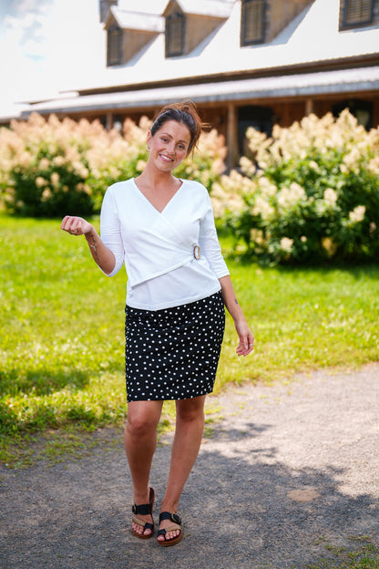 A woman wearing the Cotillon Top by Rien ne se Perd in White, a v-neck top with a wrap-over front, coconut shell buckle at the waist, and 3/4 sleeves, made from stretch cotton. She is wearing it with a polka dot skirt and standing outside. 
