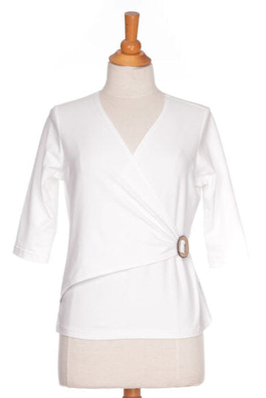 The Cotillon Top by Rien ne se Perd in White, a v-neck top with a wrap-over front, coconut shell buckle at the waist, and 3/4 sleeves, made from stretch cotton, is show on a mannequin in front of a white background. 