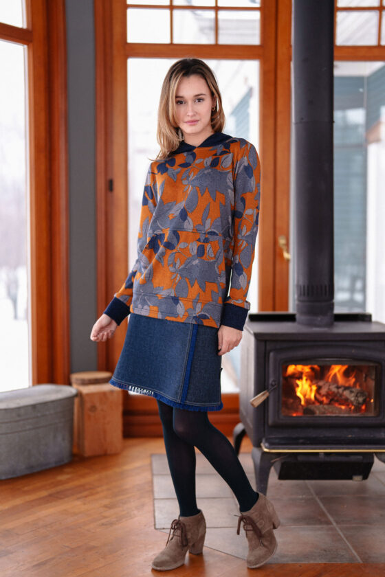 A woman wearing the Lautrec Sweater by Rien ne se Perd in Navy Foliage  with a denim skirt stands in front of a wood stove. 