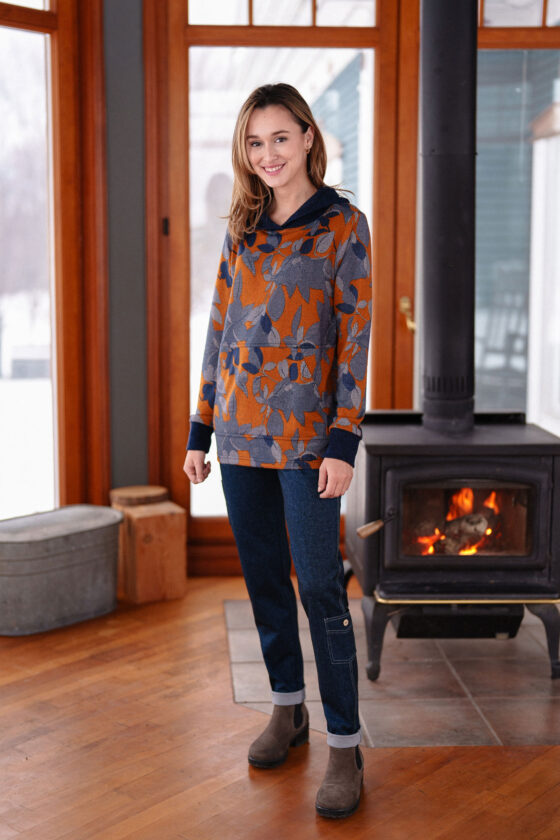 A woman wearing the Lautrec Sweater by Rien ne se Perd in Navy Foliage with jeans stands in front of a wood stove. 