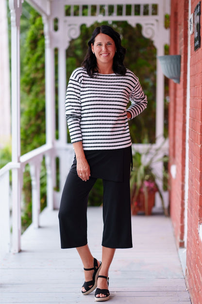 A woman wearing the Collins Reversible Sweater by Rien ne se Perd in Black/White. It's reversible back to front with stripes on side and solid fabric on the other. It has long striped sleeves and a round neck. She is wearing with black pants and standing on a porch. 