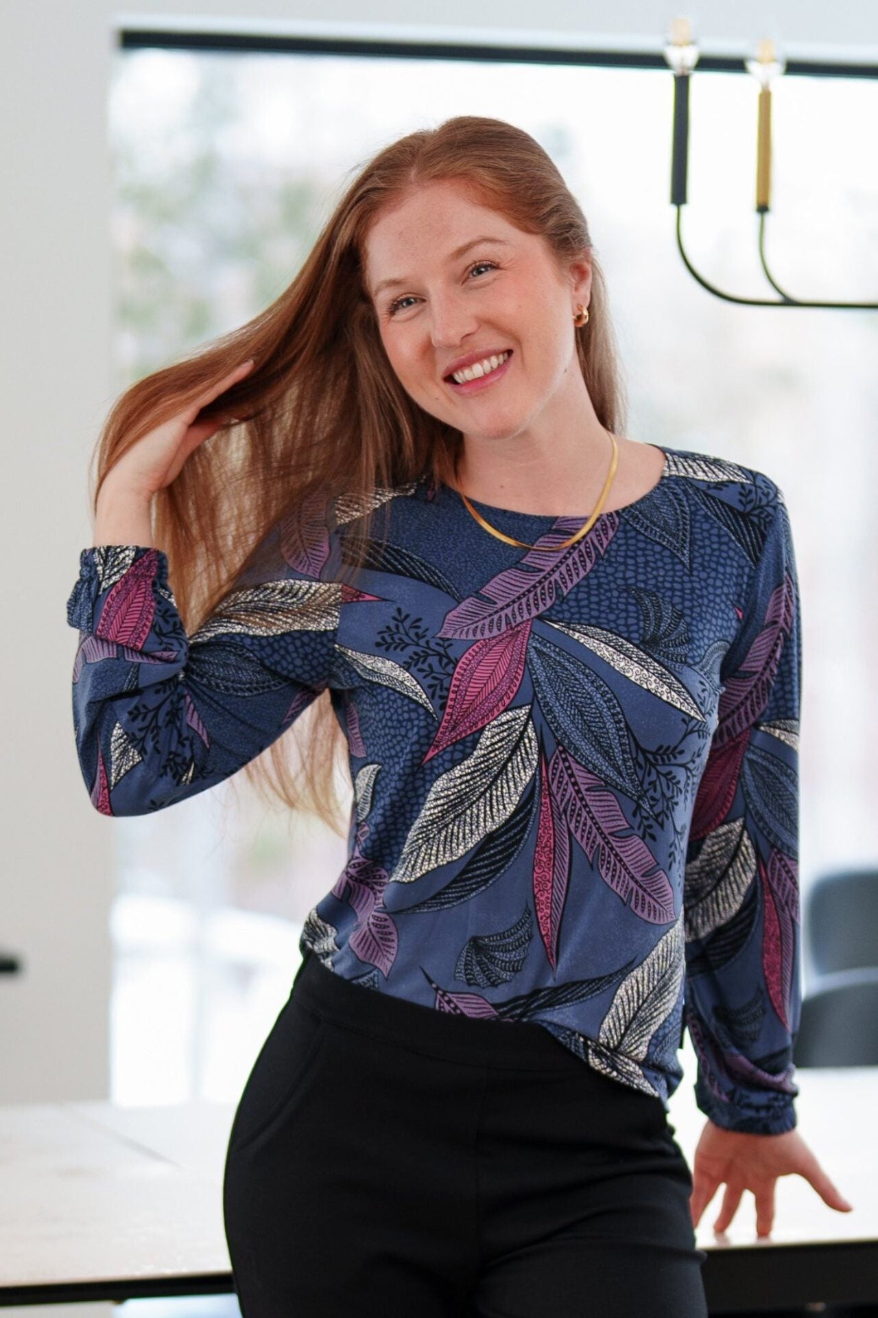 A woman wearing the St Charles Sweater by Rien ne se Perd in Foliage, a jewel toned patterned top with long sleeves and elastic cuffs. She is wearing it with the Murphy Pants in Black and standing in a modern kitchen. 
