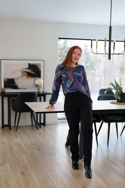 A woman wearing the St Charles Sweater by Rien ne se Perd in Foliage, a jewel toned patterned top with long sleeves and elastic cuffs. She is wearing it with the Murphy Pants in Black and 
standing in a modern kitchen. 

