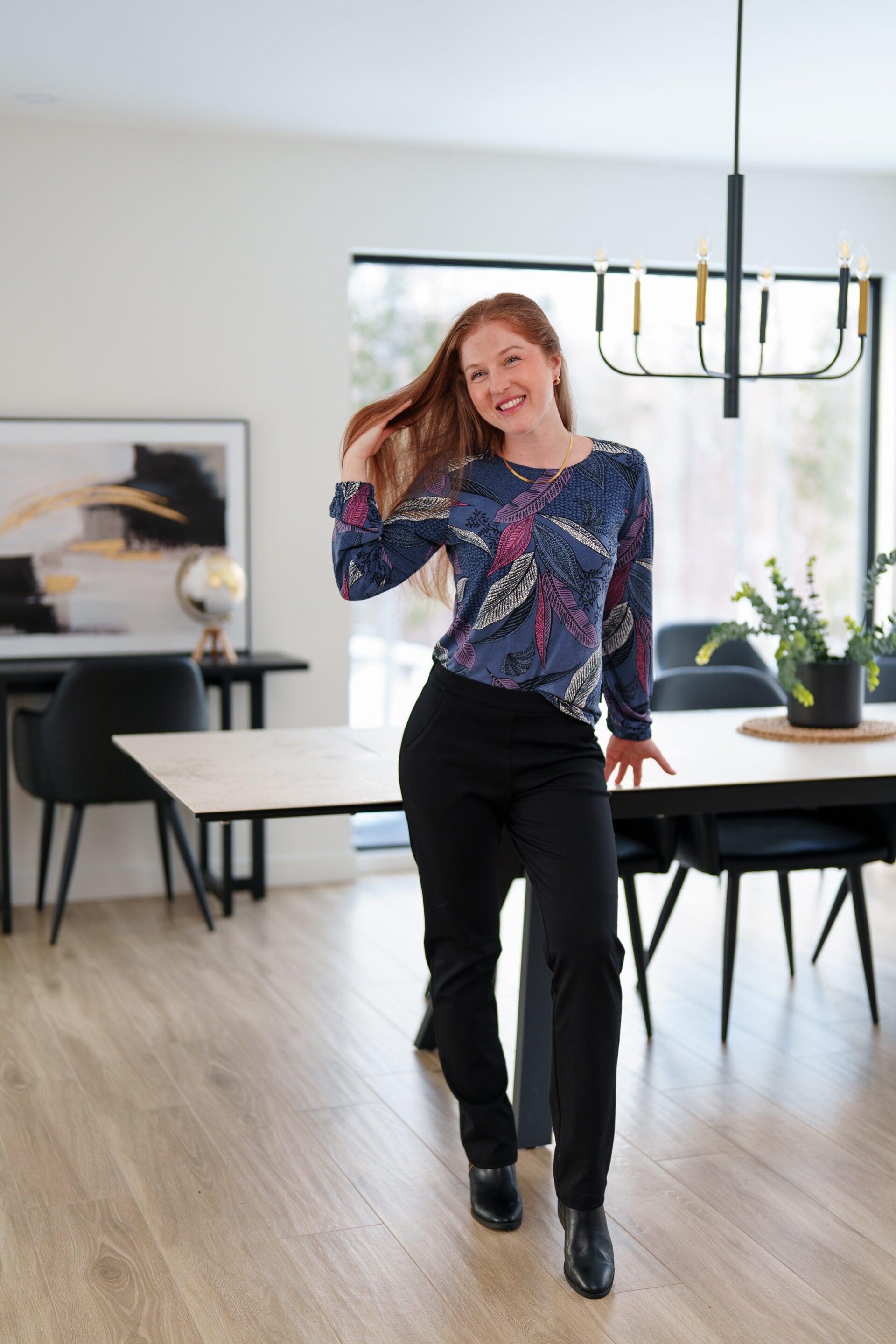 A woman wearing the St Charles Sweater by Rien ne se Perd in Foliage, a jewel toned patterned top with long sleeves and elastic cuffs. She is wearing it with the Murphy Pants in Black and standing in a modern kitchen. 
